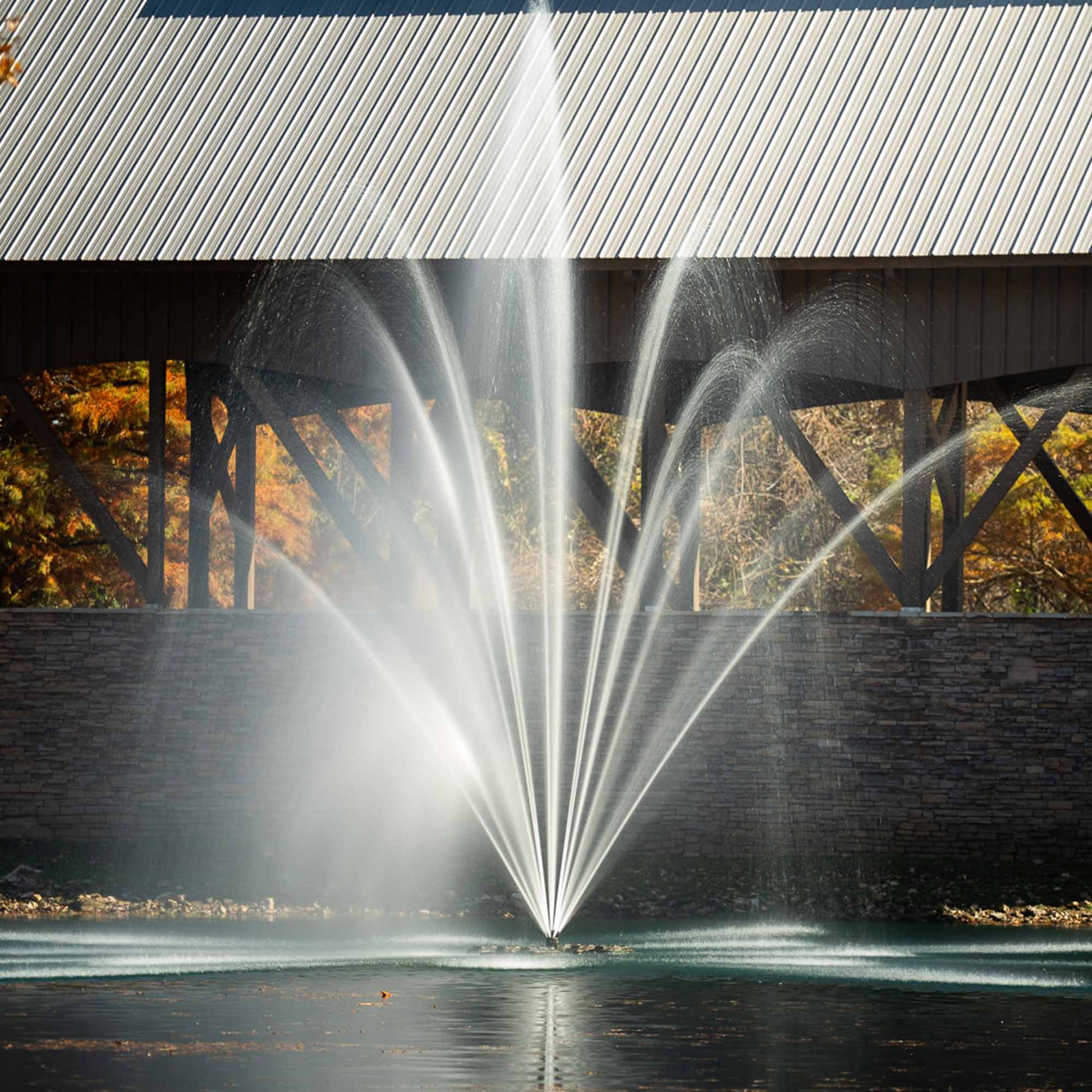 Large outdoor electric water fountain in a pond with a stone wall backdrop.