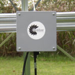 Close up of an Aeration Control Box. The box is silver and has the OWS logo on it. It is attached to a silver metal bar. There are plants in the background.