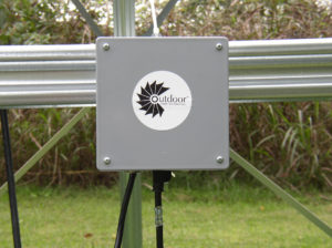 Close up of an Aeration Control Box. The box is silver and has the OWS logo on it. It is attached to a silver metal bar. There are plants in the background.