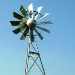 A windmill head on a single support against a blue sky with trees in the background.