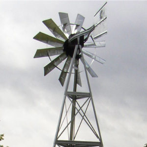 A windmill head on a single support against a blue sky with trees in the background.