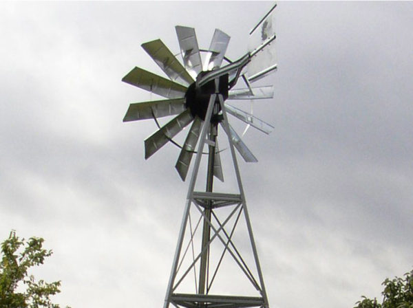 A windmill head on a single support against a blue sky with trees in the background.
