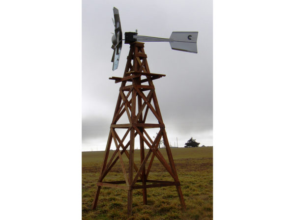 A silver windmill head on a wood base in a field with a cloudy sky.