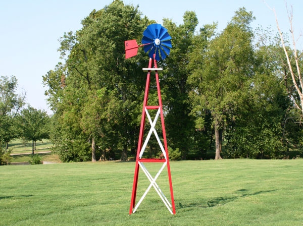 A blue, red, and white Small Backyard Windmill on grass with trees in the background.