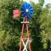 Close up of a blue, red, and white Small Backyard Windmill on grass with trees in the background.