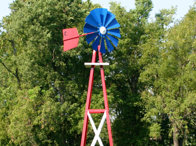 Close up of a blue, red, and white Small Backyard Windmill on grass with trees in the background.