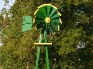 Close up of a green and yellow Small Backyard Windmill on grass with trees in the background.