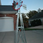 Small backyard windmill in silver with red tips. It is in a driveway next to a house.