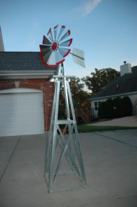 Small backyard windmill in silver with red tips. It is in a driveway next to a house.