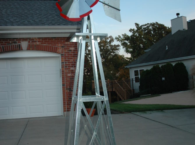 Small backyard windmill in silver with red tips. It is in a driveway next to a house.