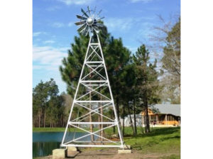 A galvanized ornamental 4-legged windmill with trees and a pond in the background.