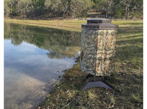 A camo fish feeder on the bank of a pond. There are trees in the background.