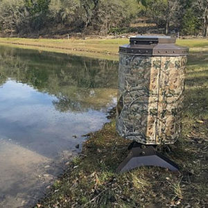 A camo fish feeder on the bank of a pond. There are trees in the background.