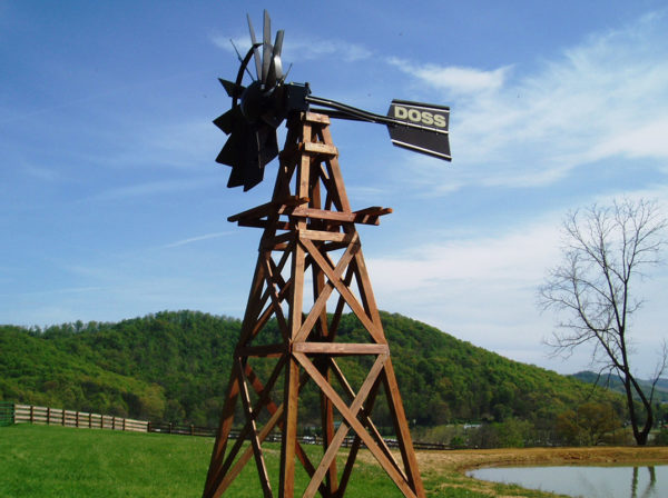 A wooden windmill with a bonze head against a background with trees, hills, and a blue sky.