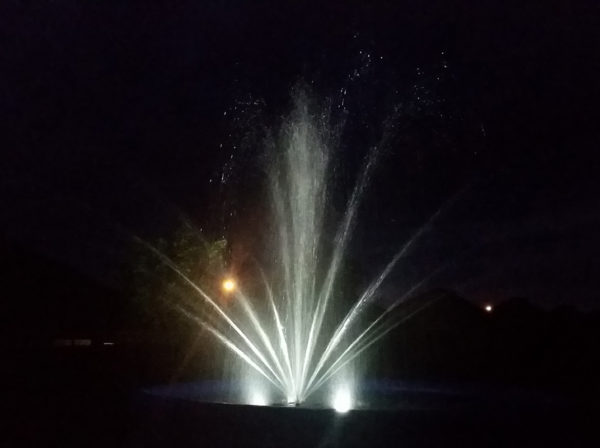 A floating pond fountain at night. It is lit up with LEDs so the water is visible in the dark.
