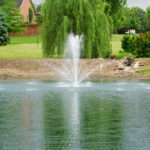 Floating pond fountain in water. The fountain sprays several streams of water into the air. The water is very blue in the foreground and less blue in the background. In the background there are trees and buildings.