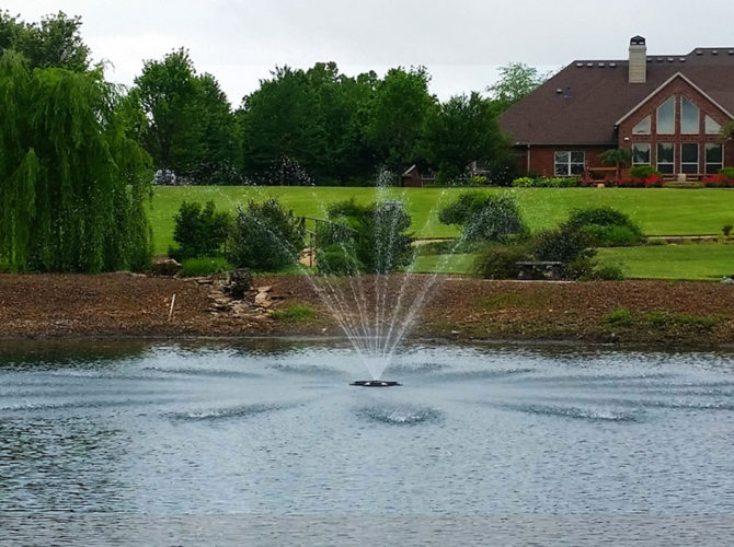Floating pond fountain in water. The fountain sprays several streams of water into the air. The water is very blue in the foreground and less blue in the background. In the background there are trees and buildings.