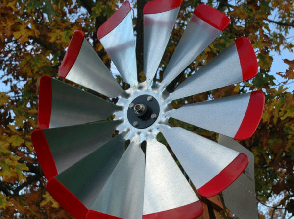 Close up on windmill head in silver with red tips on a wooden windmill body. In the background there are trees.