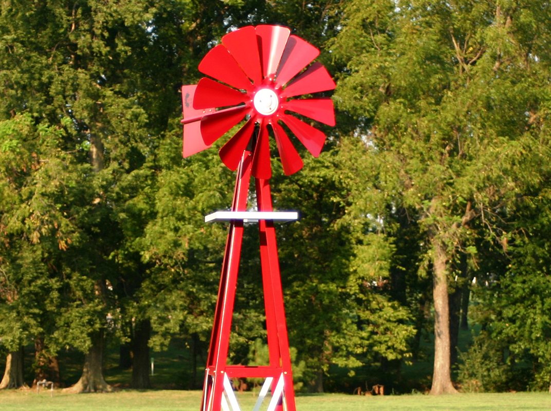 Close up of a red and white Small Backyard Windmill on grass with trees in the background.
