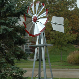 A windmill head in silver with red tips. In the background are trees and a house.