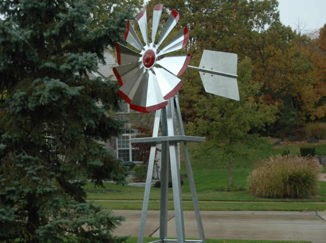A windmill head in silver with red tips. In the background are trees and a house.