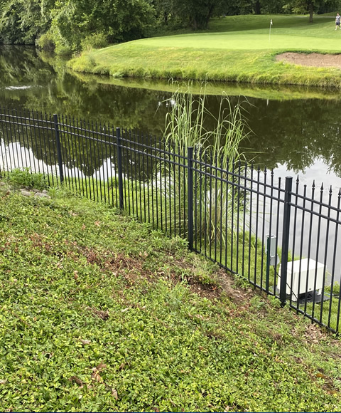 There is a fence in the foreground. In the background is a pond, tan aeration cabinet and green grass.
